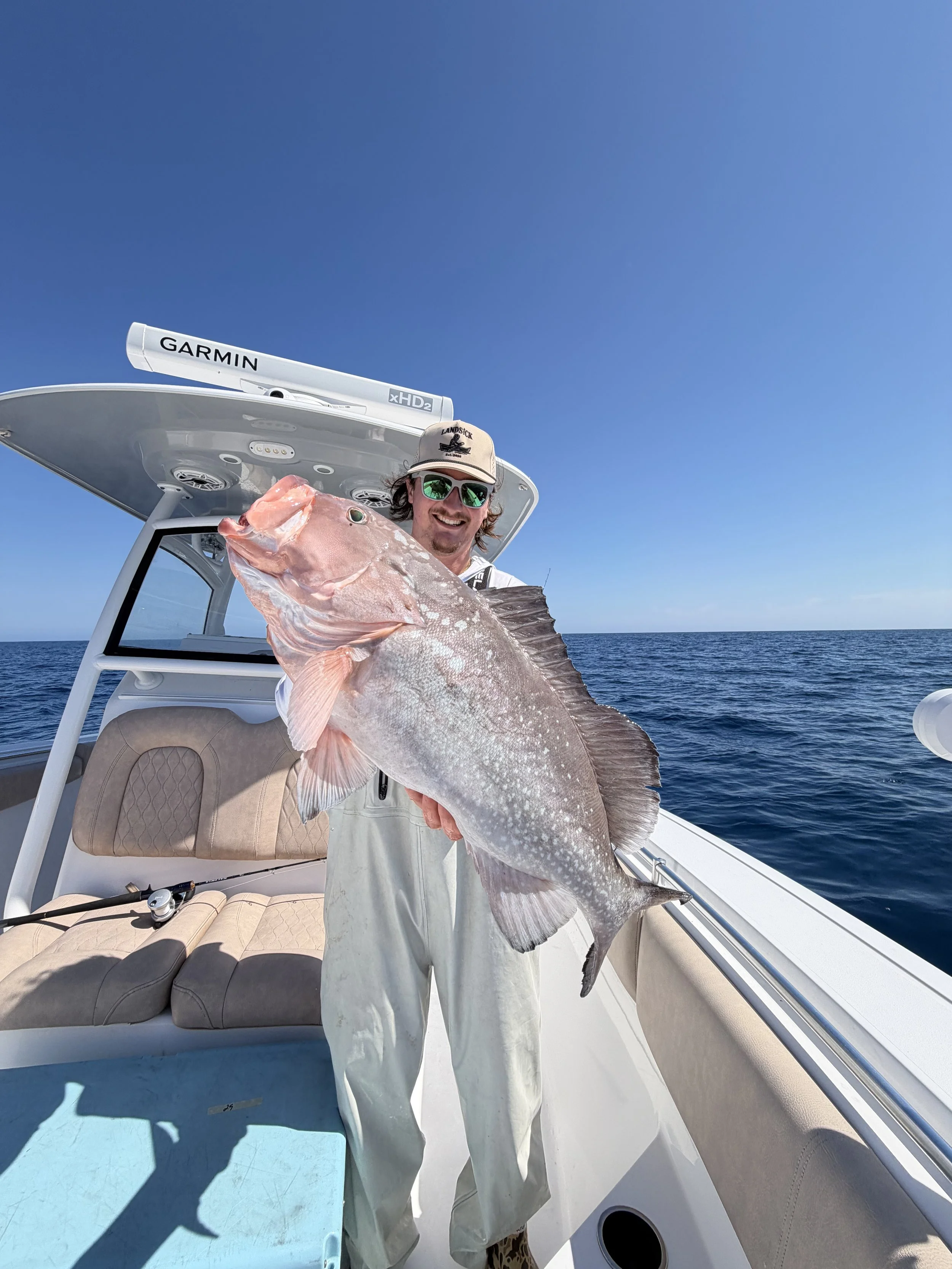 Man kneeling on boat deck, holding two large fish, smiling, wearing a black cap, sunglasses, white hoodie, and shorts, with water and houses in the background.