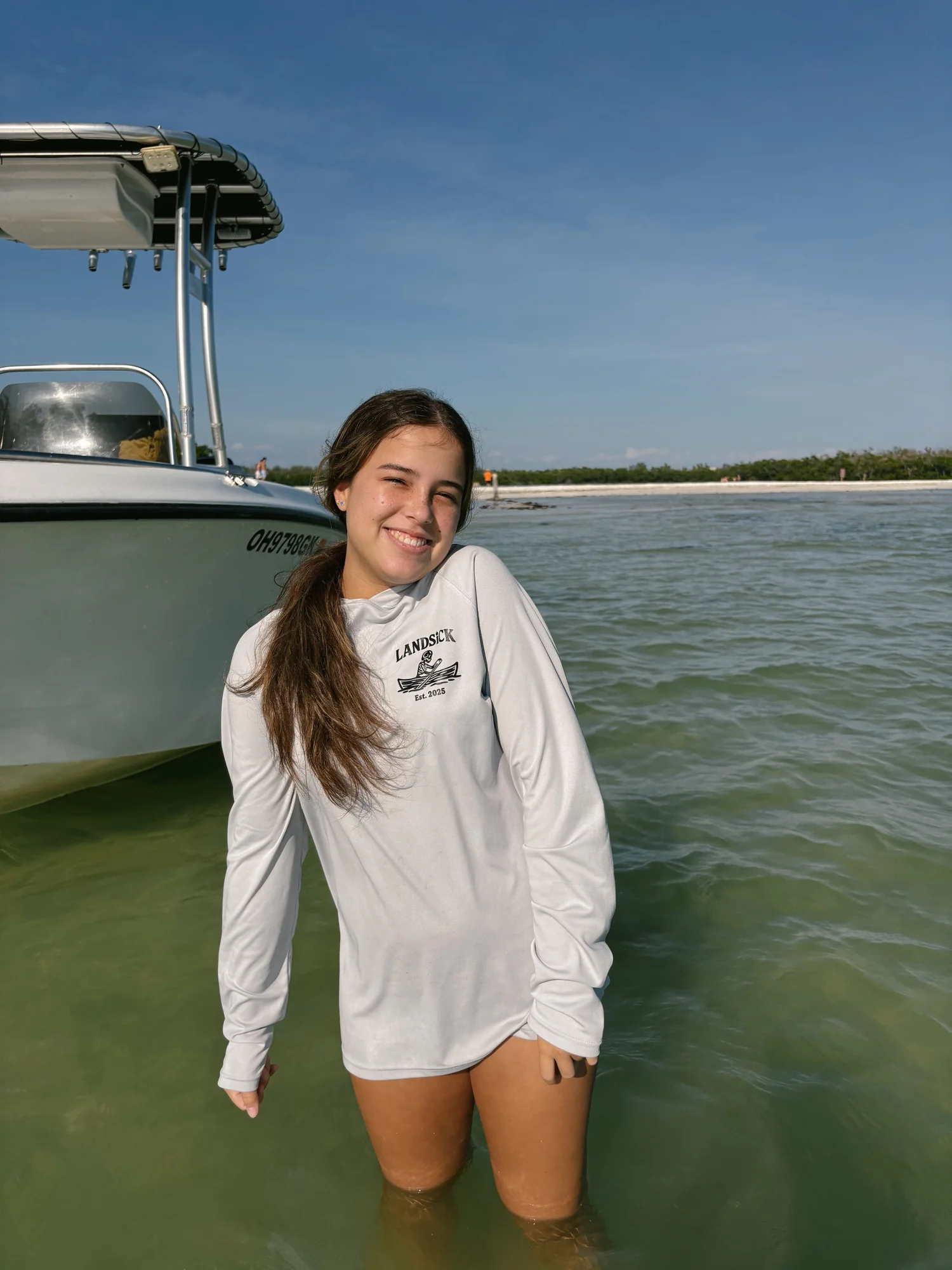 A girl standing knee-deep in water next to a boat on a beach with a clear blue sky.