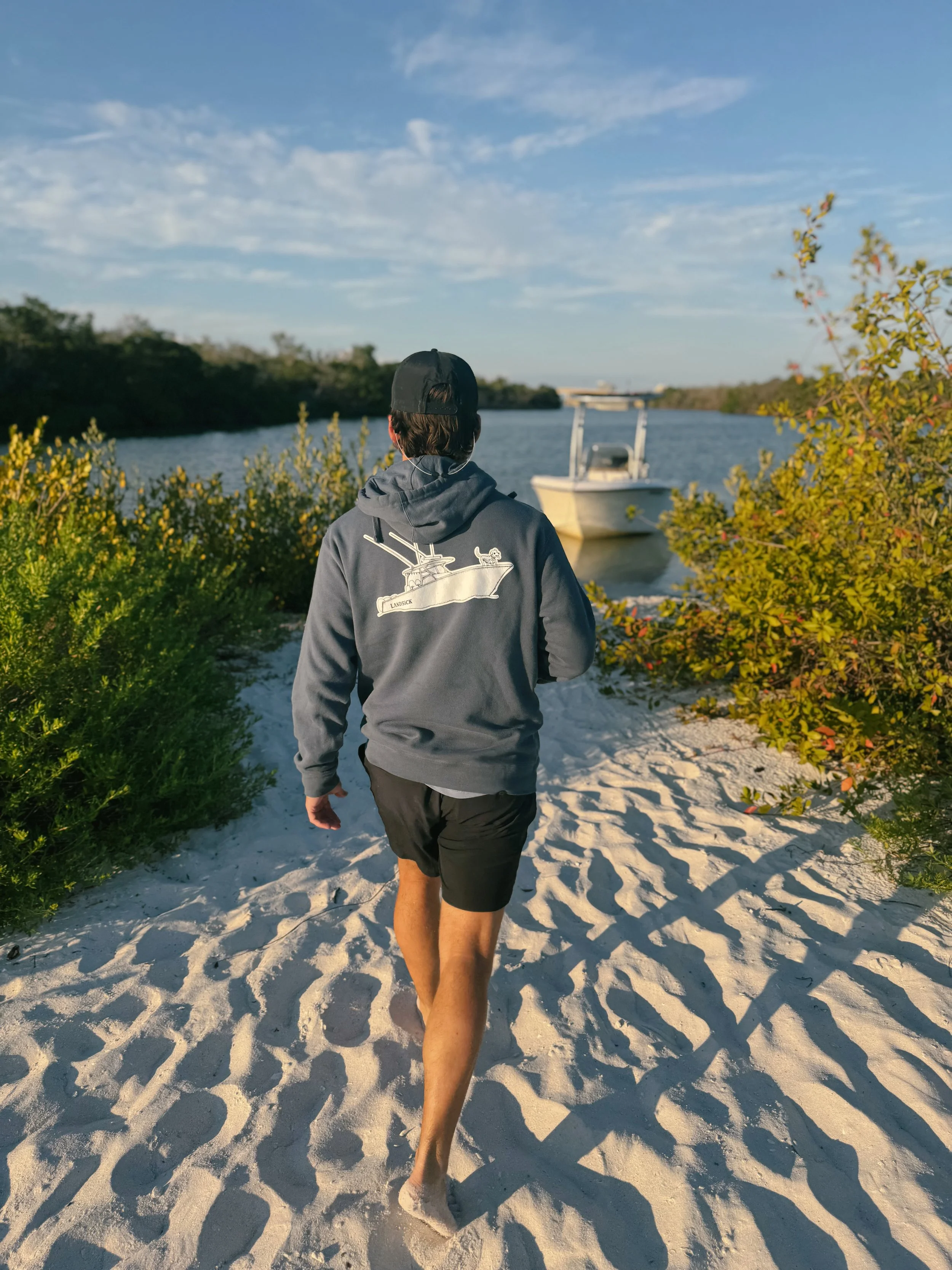 A man in a LANDSICK hoodie walks down a sandy path toward a boat on the water.