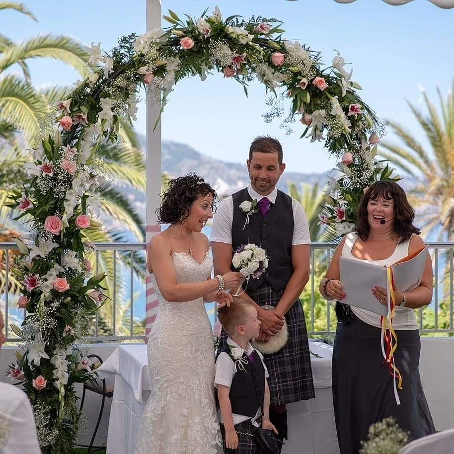 A wedding ceremony with a bride, groom, a young boy, and a woman officiating under a floral arch, overlooking palm trees and a body of water.