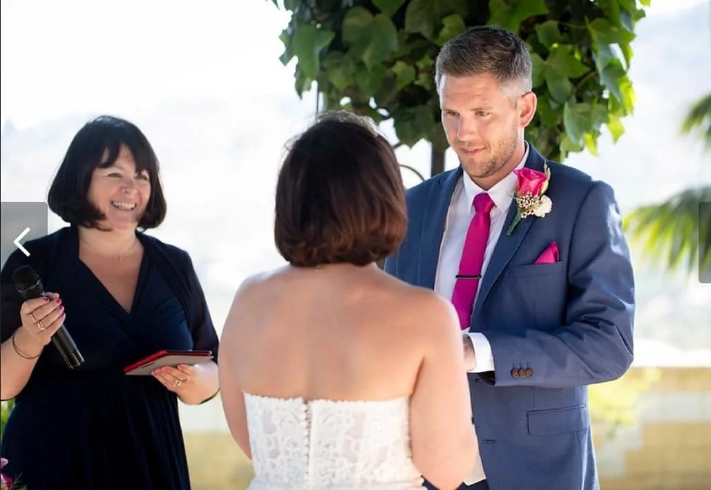 A couple exchanging wedding vows outdoors, with a female officiant holding a microphone and a notebook, under a tree on a sunny day.