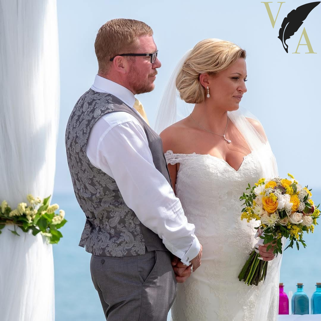 A bride and groom standing hand in hand during their outdoor wedding ceremony, with the ocean in the background. The bride is holding a bouquet of yellow and white flowers, and both appear to be emotionally focused.