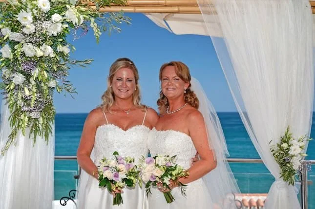 Two women in white wedding dresses holding bouquets of flowers, standing under a decorated canopy on a deck overlooking the ocean during a wedding ceremony.