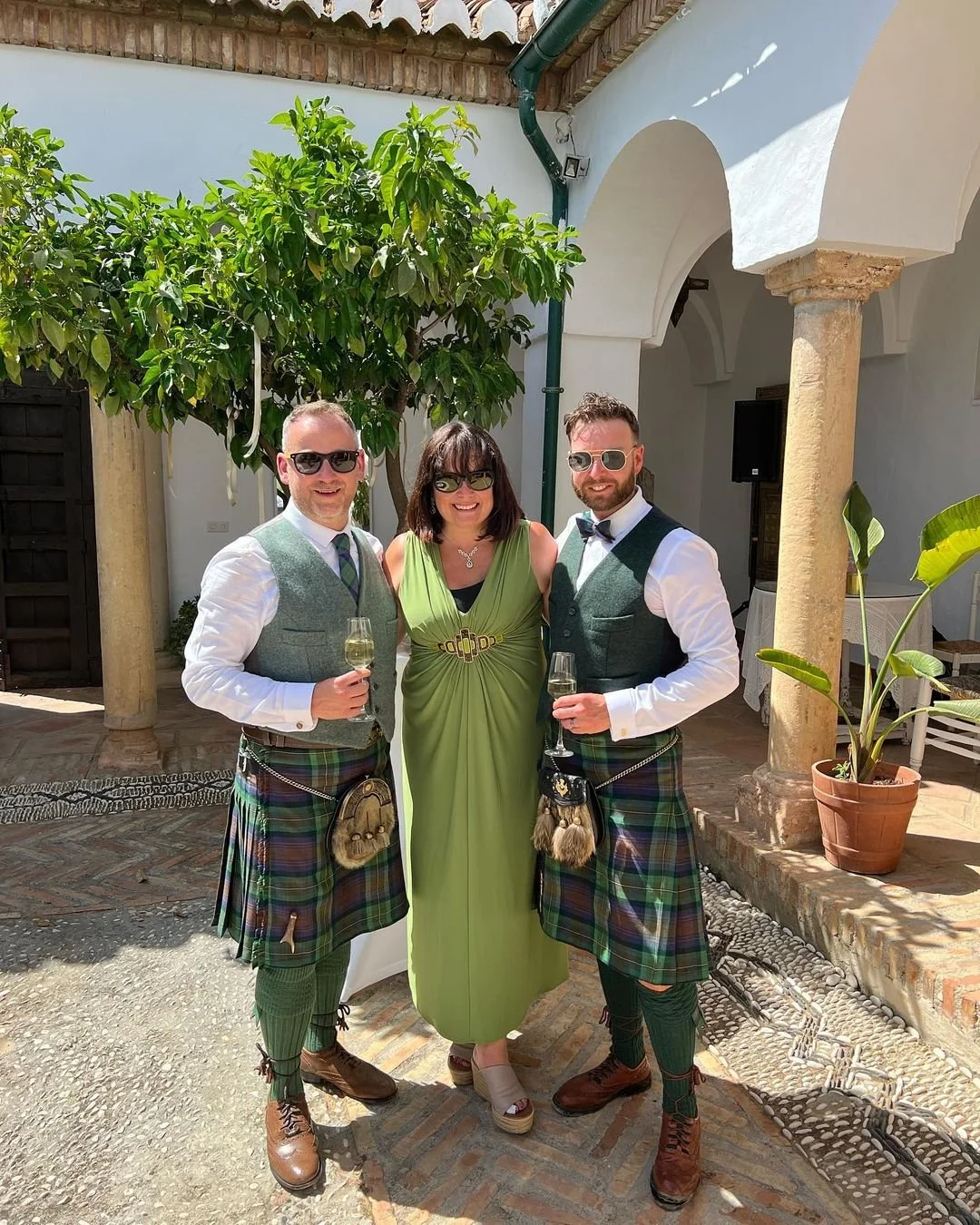 Three people, two grooms in traditional Scottish kilts and Nikki Kulin, professional wedding celebrant, standing outdoors under a tree, holding glasses of champagne, smiling. Nikki has just delivered their unique, bespoke wedding ceremony.
