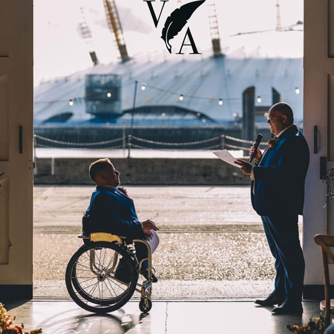A man in a suit reading from a paper with a microphone to a young boy in a wheelchair, standing at an open doorway with a cityscape and bridge in the background.