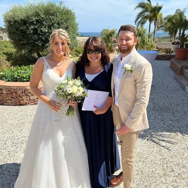 A bride in a white wedding dress holding a bouquet, standing next to a man in a beige suit, and Nikki Kulin, professional Wedding Celebrant after having delivered their unique wedding ceremony in southern Spain.