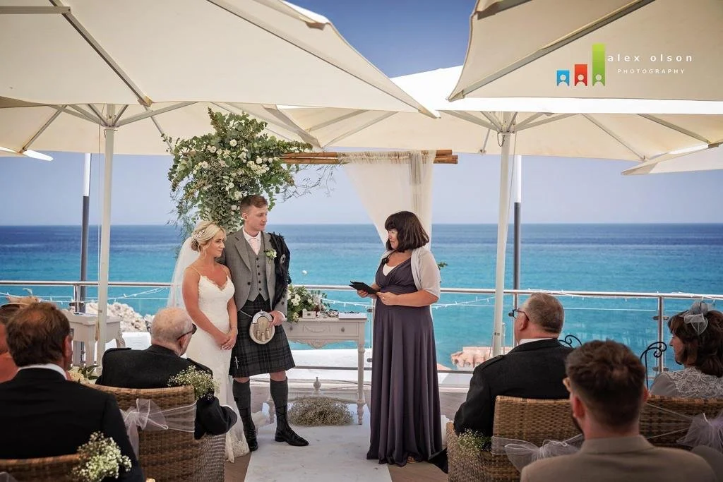 A wedding ceremony takes place outdoors on a terrace overlooking the ocean under large white umbrellas, with a bride and groom facing a woman officiant, and guests seated nearby.