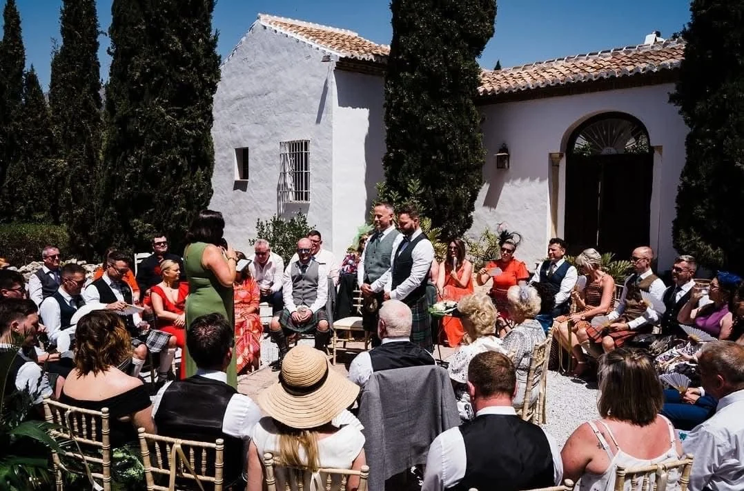 A wedding ceremony outdoors with guests seated around, a woman in green officiating, and two men standing as grooms, in front of a white building with trees and blue sky.