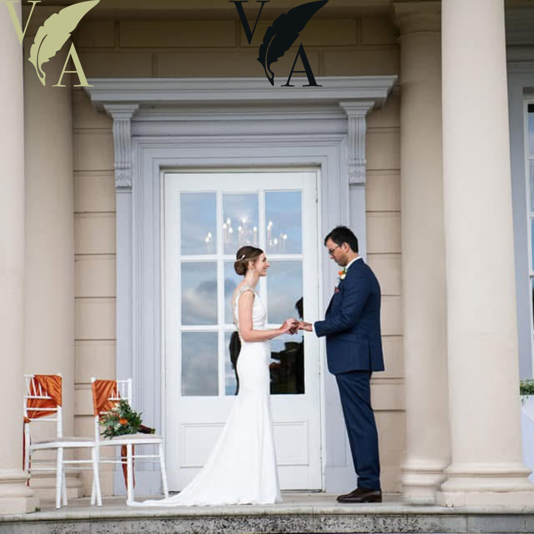 A bride and groom exchanging vows outside a building with white columns and large window, chairs with orange ribbons and floral arrangements nearby.