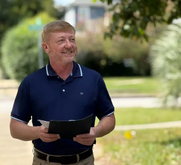 Chris Stephens, A middle-aged man with light brown hair and a beard, smiling and holding a black folder or notebook, standing outdoors in a park with trees and green grass.