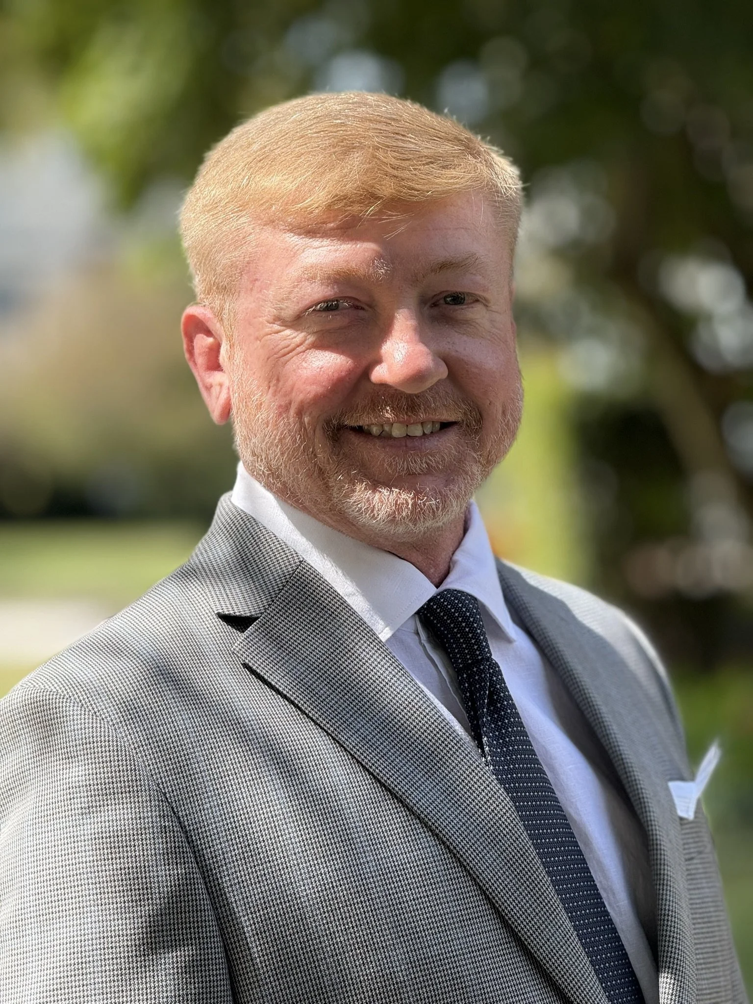 A  picture of Chris Stephens smiling with red hair and a beard wearing a light gray suit, white shirt, and dark tie, outdoors with blurred greenery in the background.