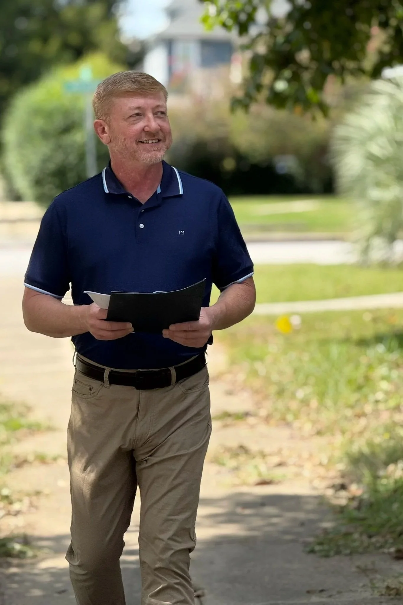 Chris Stephens, A middle-aged man with light hair and a beard walking outdoors on a sunny day, holding a black folder, dressed in beige pants and a navy polo shirt with white trim, smiling with trees and a building in the background.