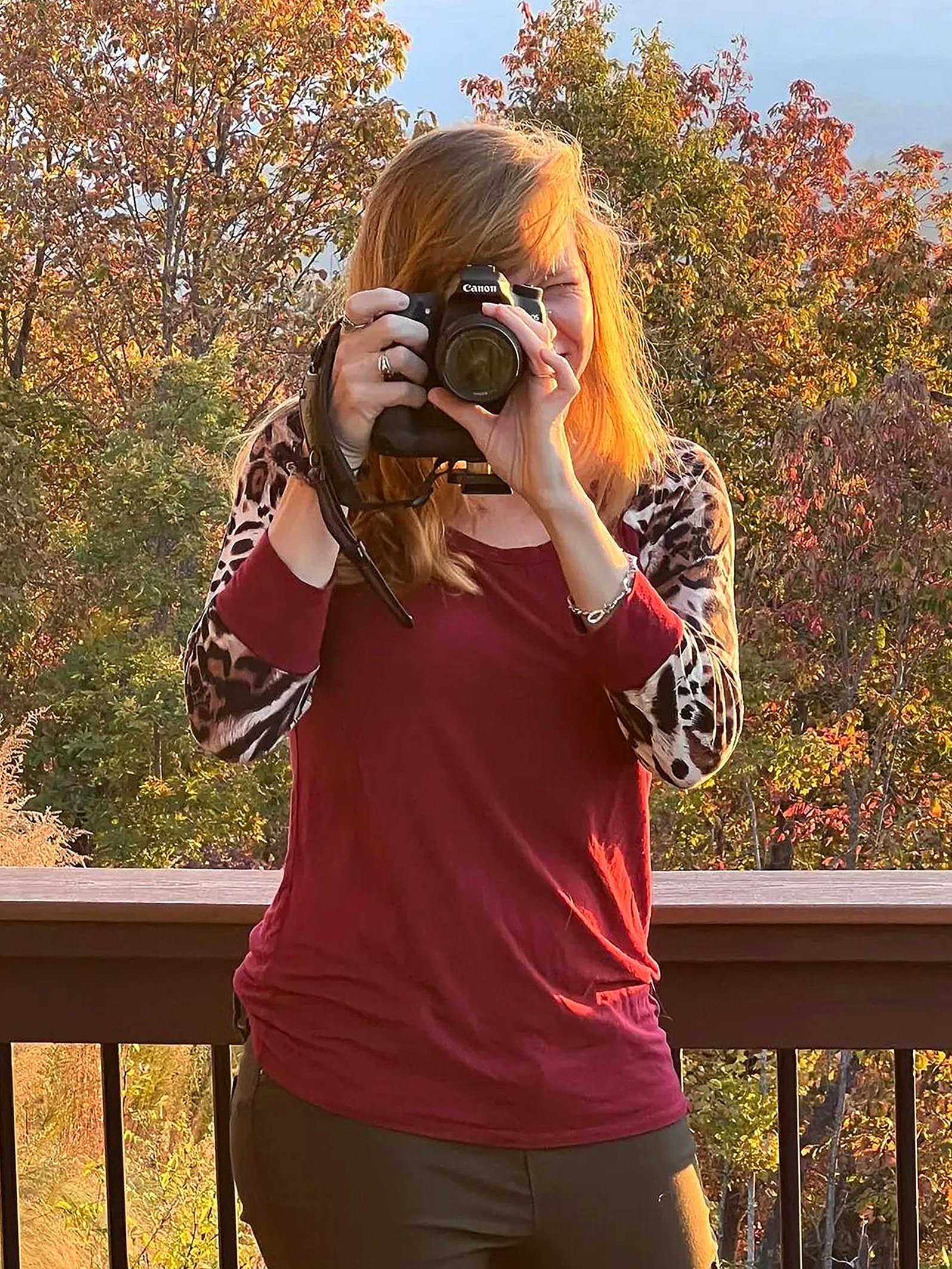 A woman with red hair taking a photo outdoors during autumn, with colorful fall trees in the background.