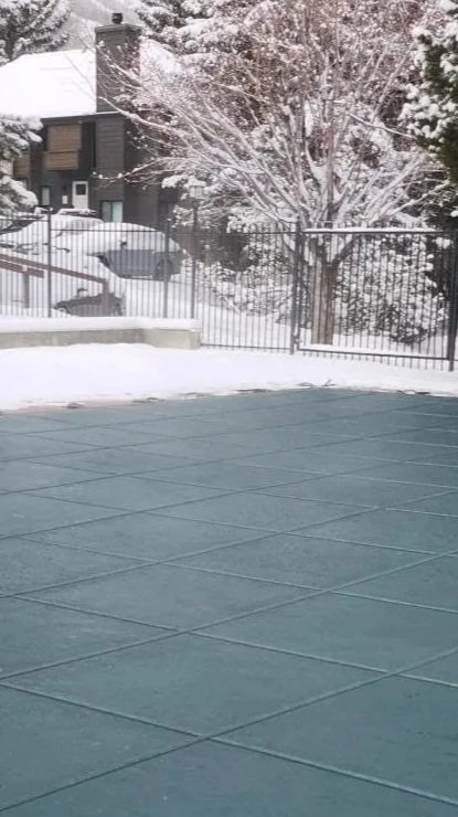 Outdoor swimming pool covered with safety cover. Surrounded by a snow-covered patio with a fence, trees, and houses in the background.