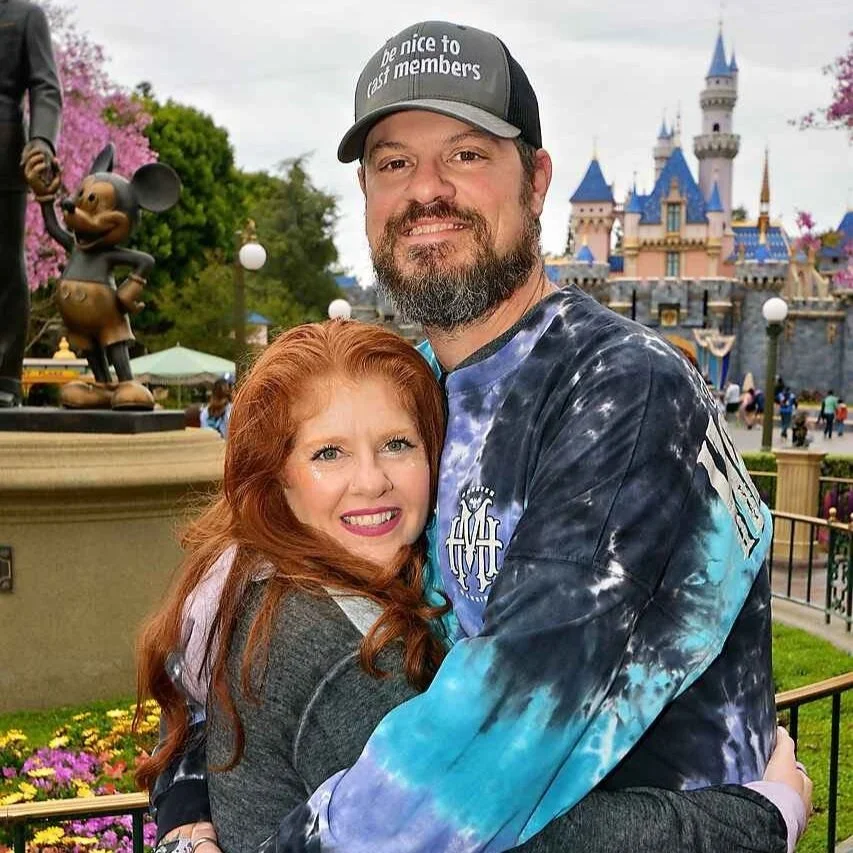 A smiling man and woman hugging in front of Sleeping Beauty Castle at Disneyland, with a Mickey Mouse statue nearby.
