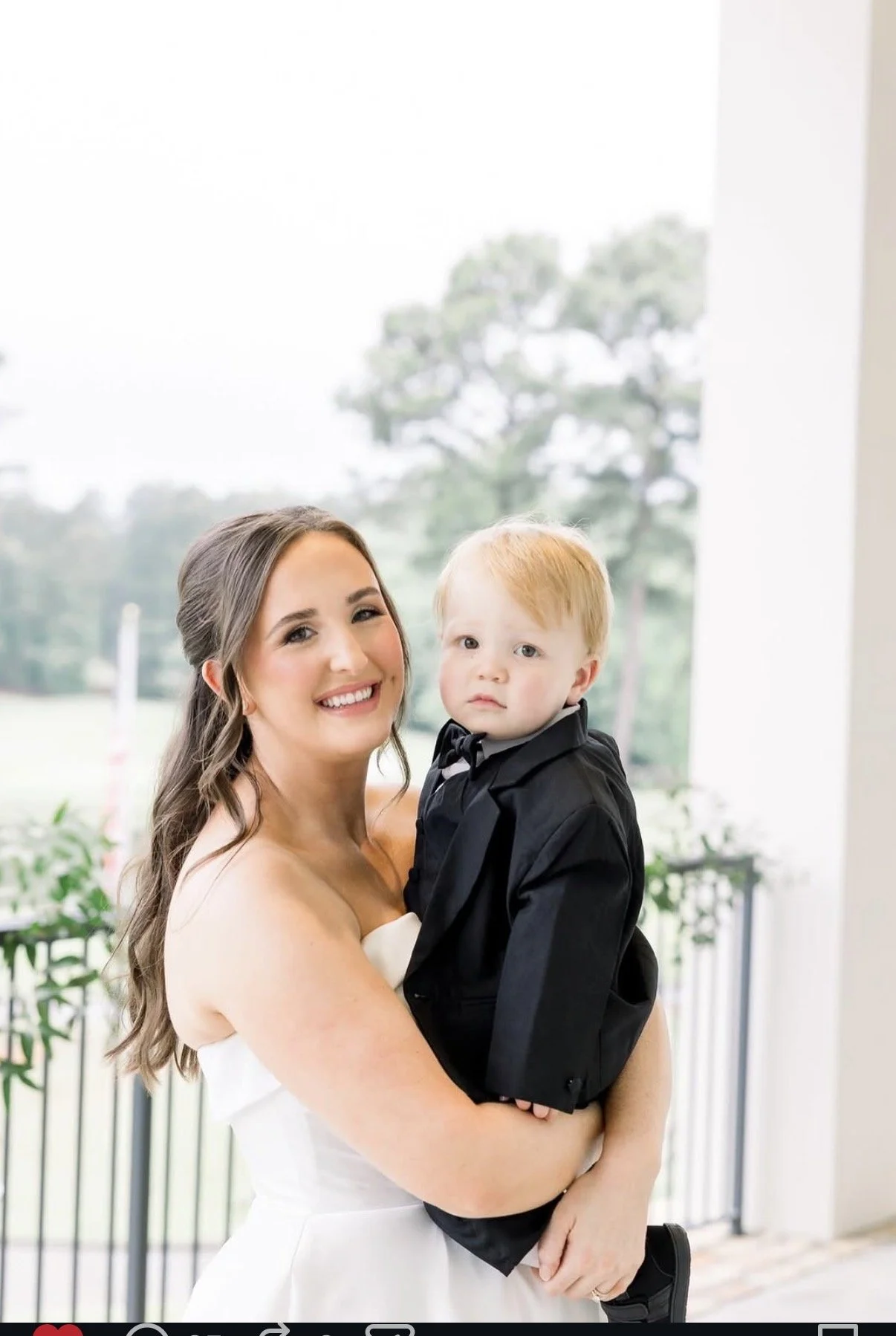A smiling woman in a white dress holding a young boy dressed in a black tuxedo with a bow tie, standing outside near a white building with trees visible in the background.
