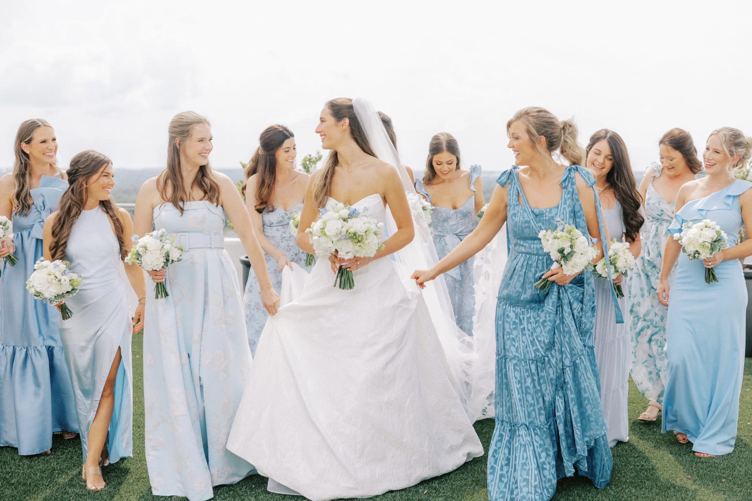 Bride surrounded by bridesmaids in blue dresses holding bouquets outdoors on a cloudy day.