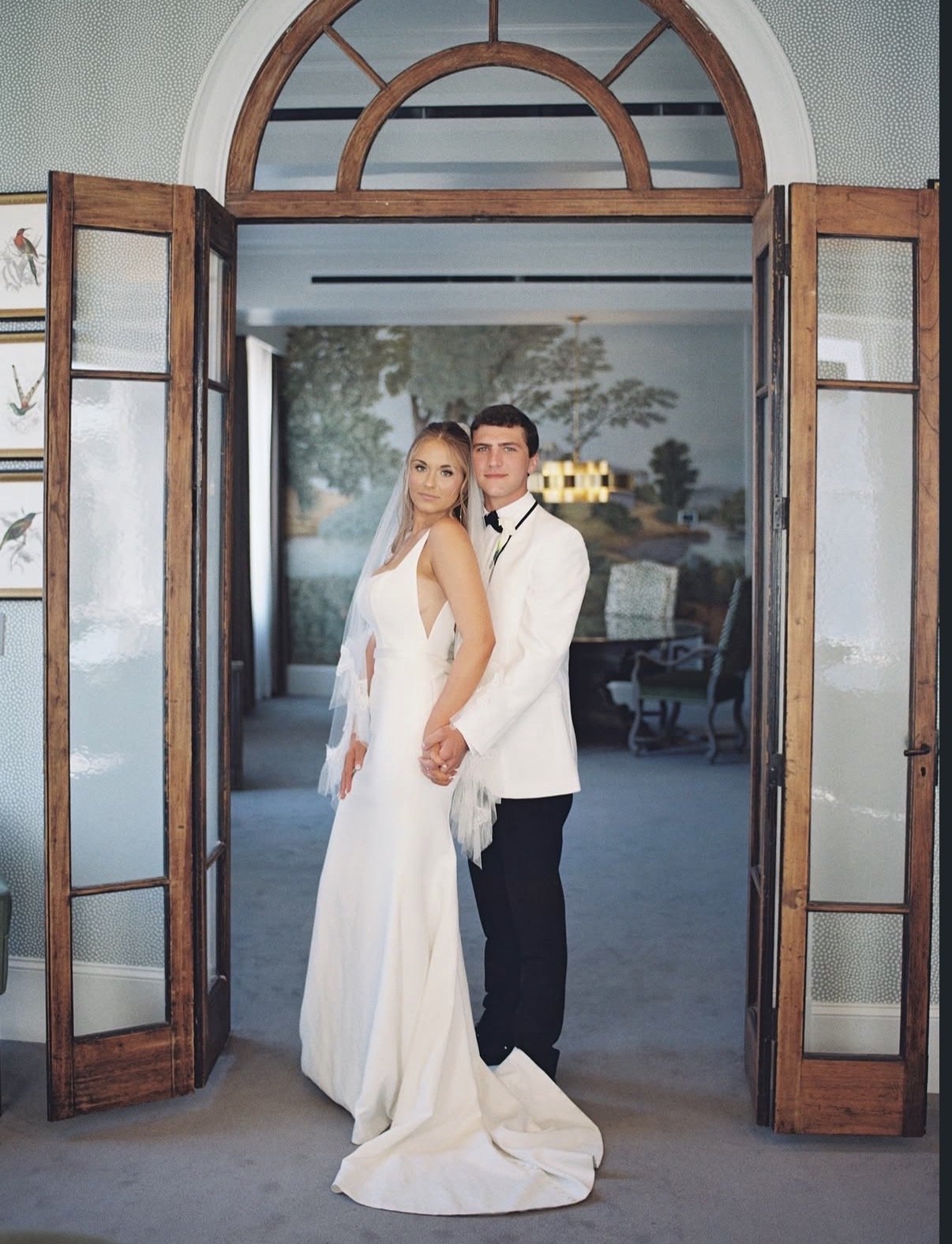 A bride and groom standing hand in hand in front of open wooden and glass double doors. The bride is wearing a long white wedding dress with a veil, and the groom is dressed in a white tuxedo jacket with black pants. They are indoors, with a view of 