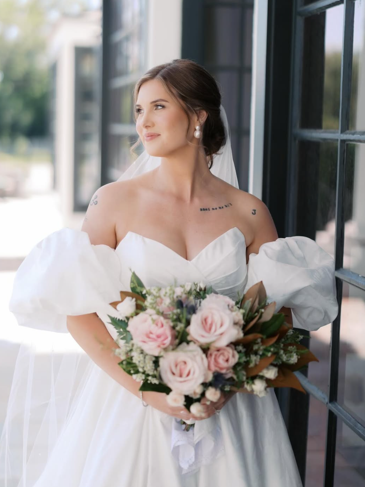 Bride in white wedding gown holding a bouquet of pink and white flowers, standing near a window with black framing, looking to the side with a gentle smile.