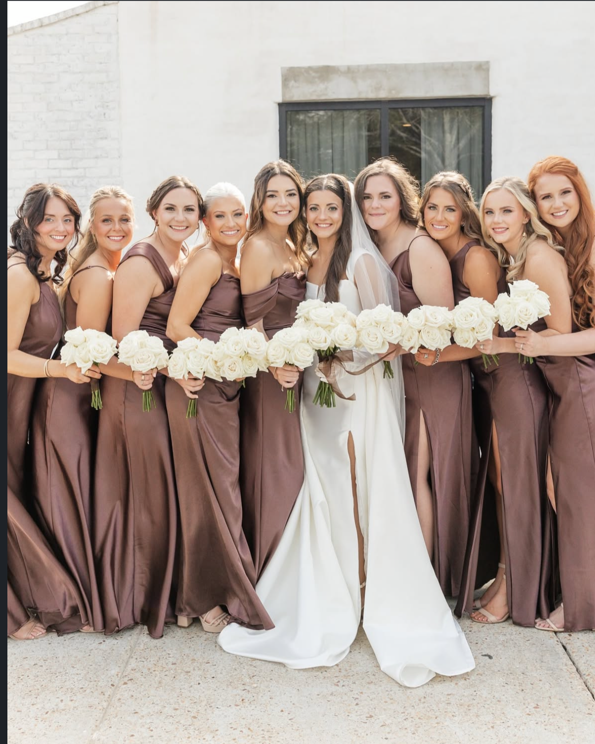 Group of women in matching brown bridesmaid dresses holding white roses with a bride in a white wedding gown and veil, standing outdoors in front of a white wall.