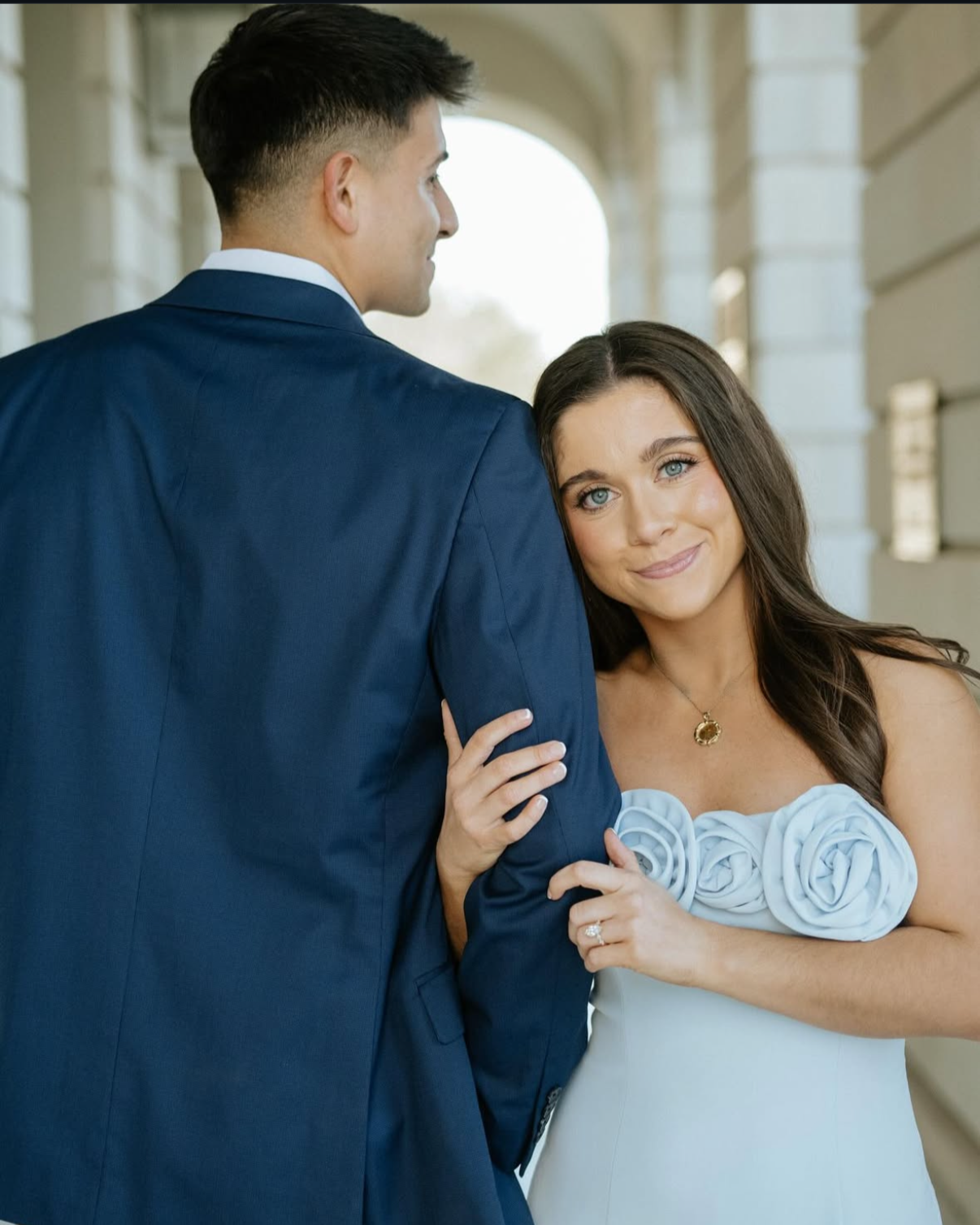 A woman in a white dress with floral accents and a man in a navy suit posing in front of a stone building, with the woman leaning her head on the man's shoulder and smiling.
