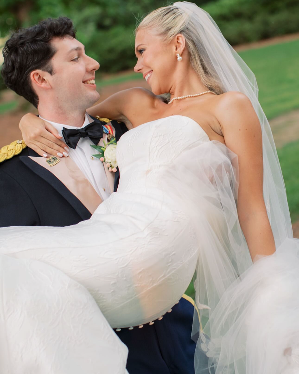 A bride and groom share a joyful moment outdoors. The groom is dressed in a formal military uniform with medals, and the bride is in a white wedding gown with a veil, pearl necklace, and earrings. The groom is holding the bride in his arms as they sm