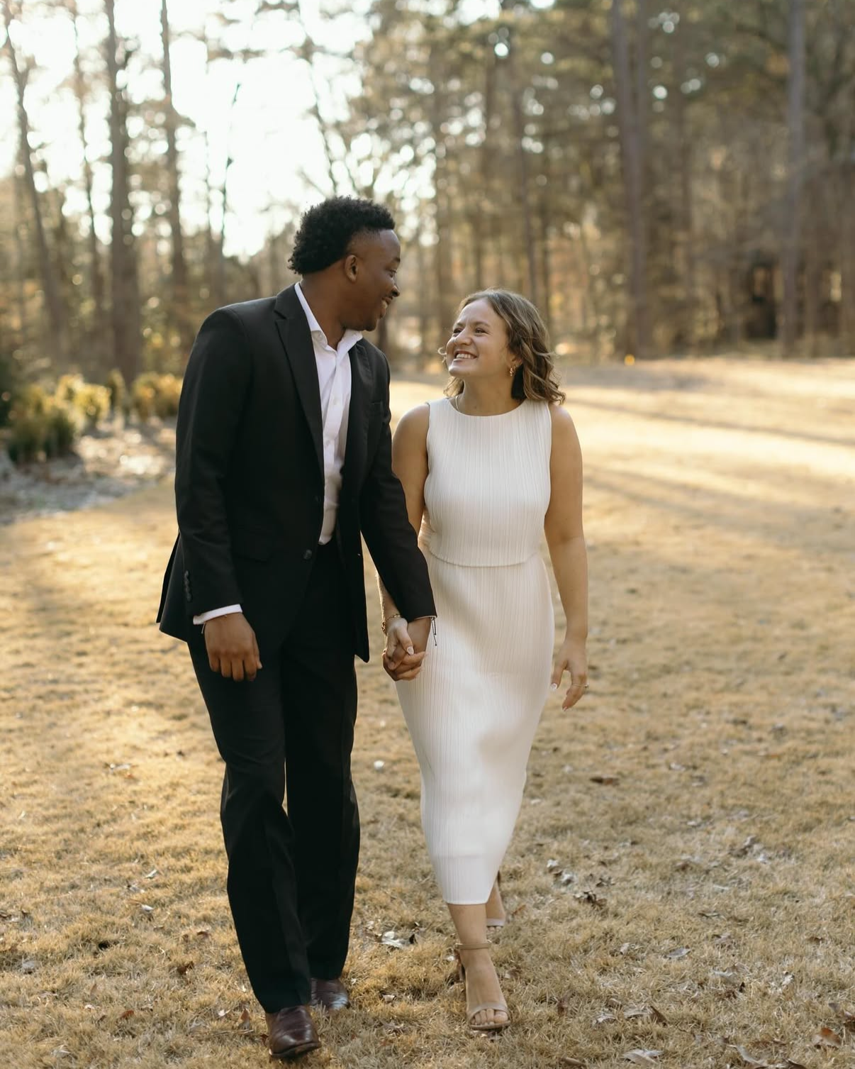 A couple walking hand in hand outdoors in a wooded area, smiling and looking at each other. The man is wearing a black suit and white shirt, and the woman is wearing a white sleeveless dress and heels.