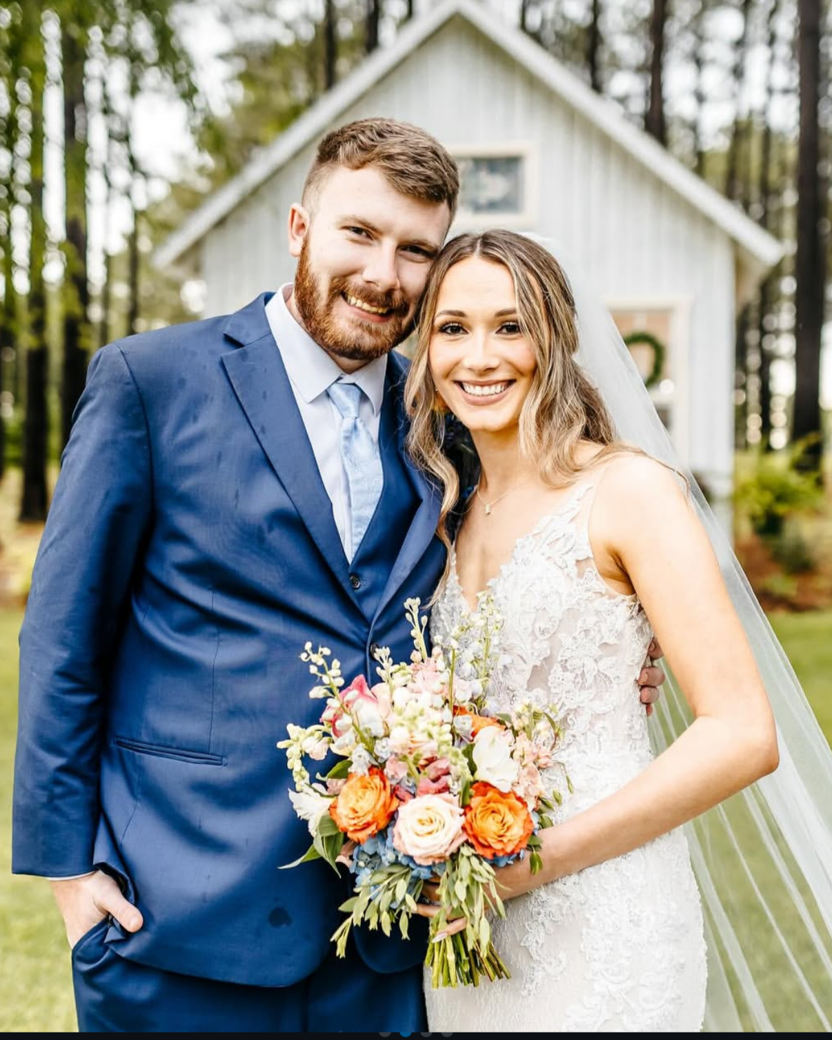 A smiling newlywed couple stands outdoors, the groom wearing a blue suit and the bride in a lace wedding dress holding a colorful bouquet of flowers, with a small white house or chapel in the background surrounded by trees.