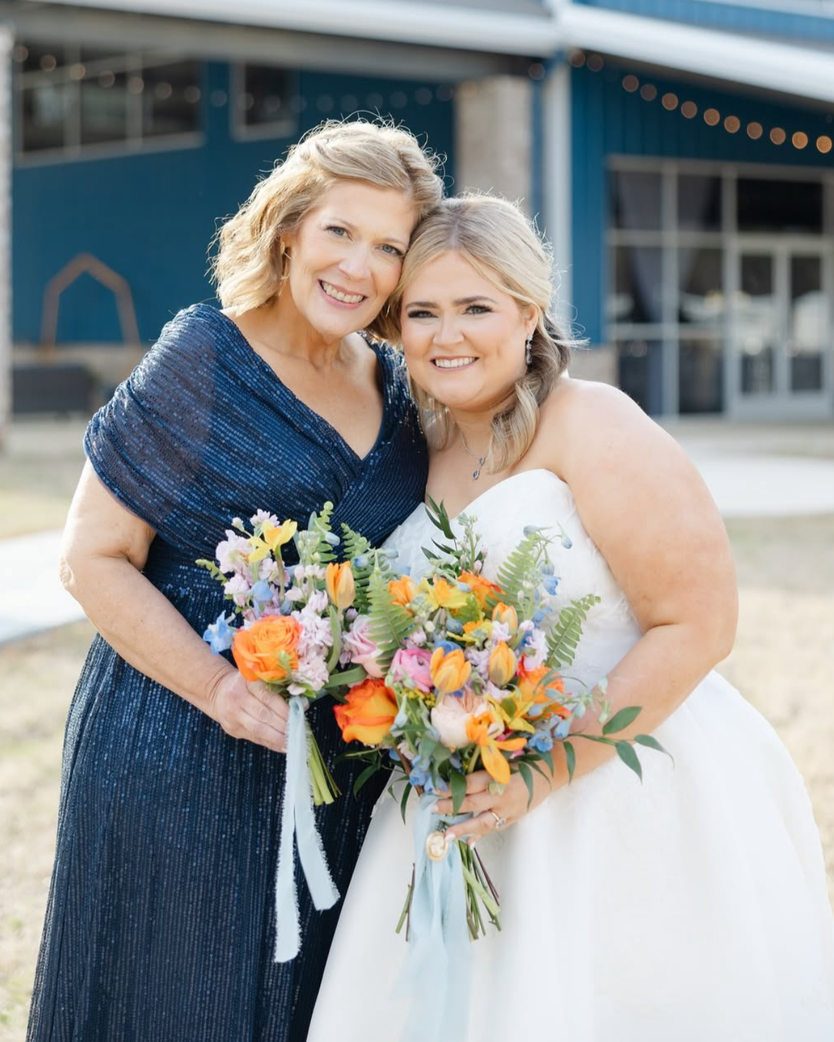 A woman in a white wedding dress and a woman in a dark blue dress smiling and holding colorful bouquets outside a building.