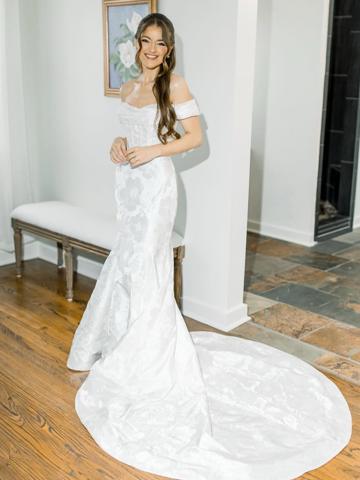 A woman in a white wedding gown smiling, standing indoors on a wooden floor next to a white bench with a painting on the wall behind her.