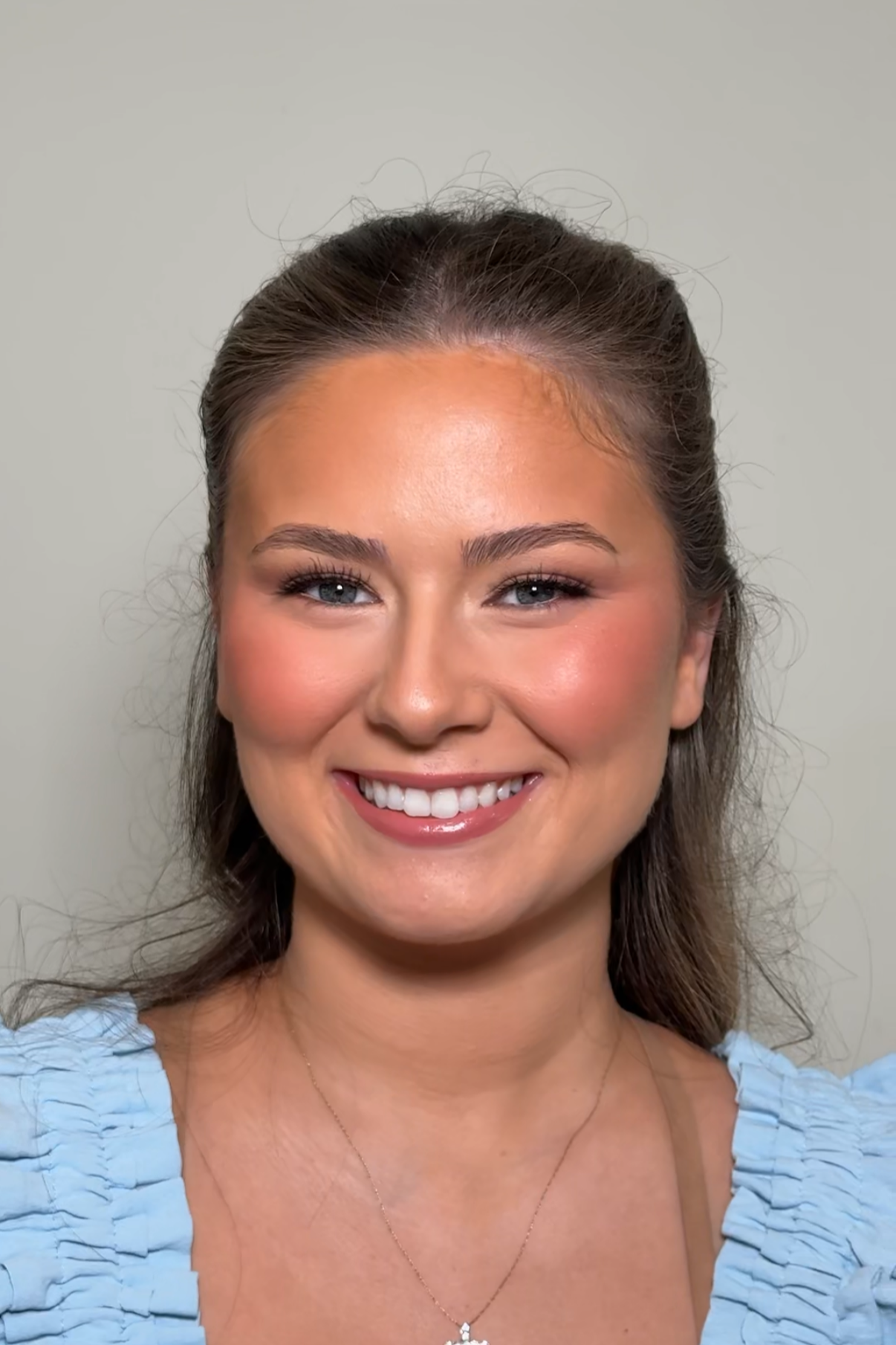 A woman smiling with braces, wearing a light blue top and a necklace, against a plain background.