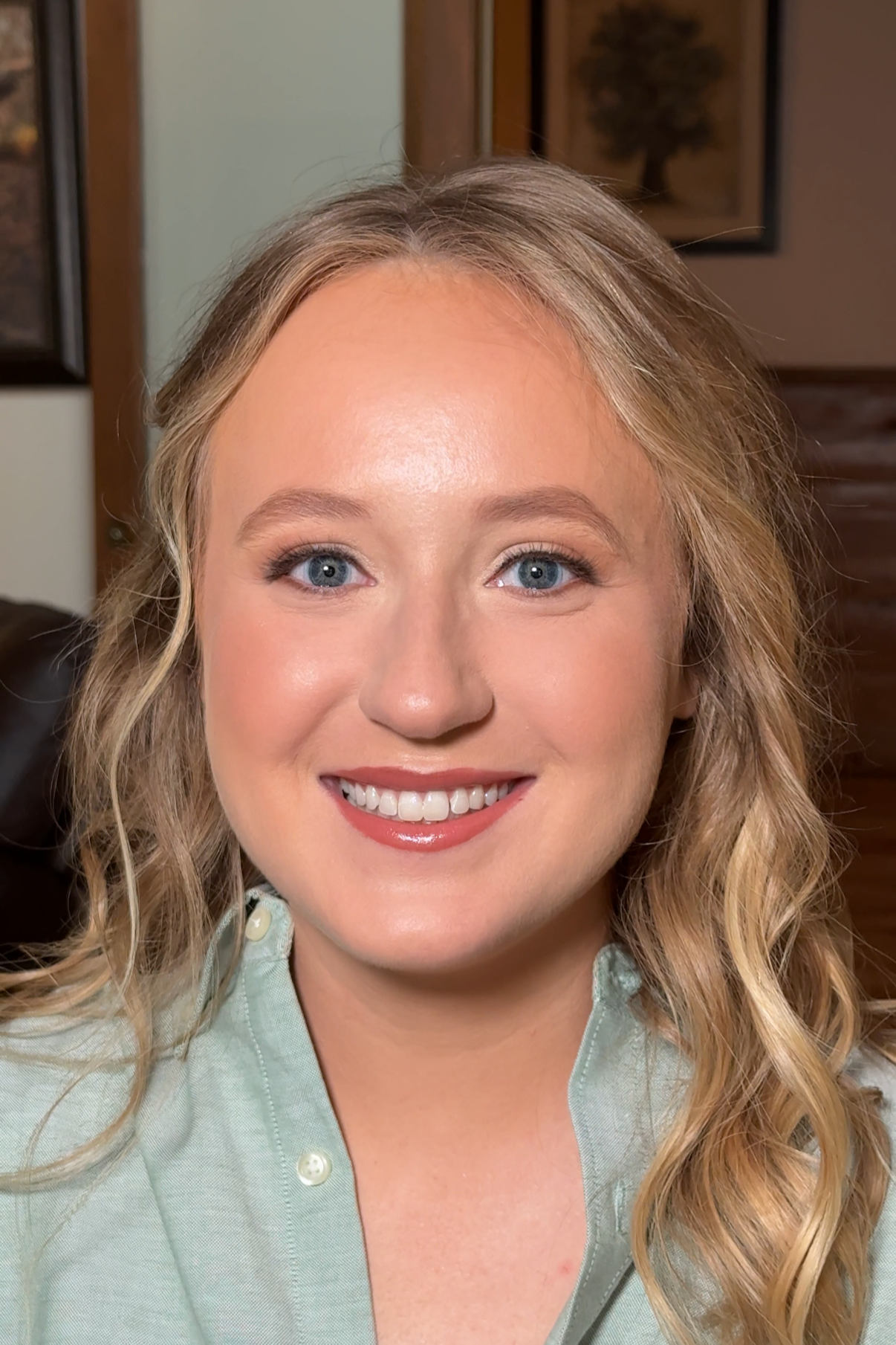 Close-up portrait of a young woman with blonde curly hair, blue eyes, and a bright smile, wearing a light green shirt, in a cozy indoor setting.
