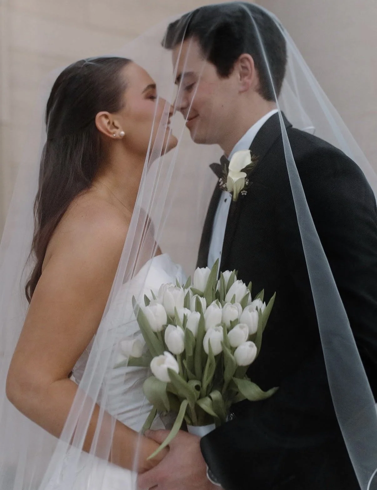 A bride and groom stand close with their foreheads touching, wearing wedding attire. The bride holds a bouquet of white tulips and is partially covered by a veil, while the groom wears a tuxedo with a boutonniere.