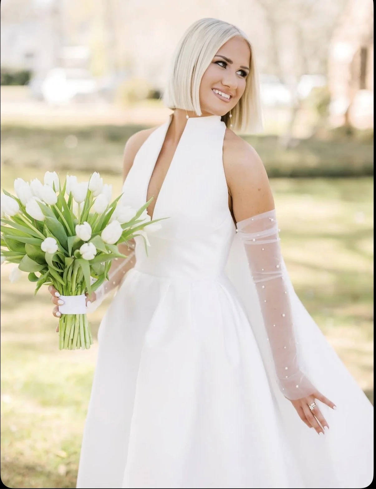 A woman in a white wedding dress holding a bouquet of white tulips outdoors, smiling and looking to the side.