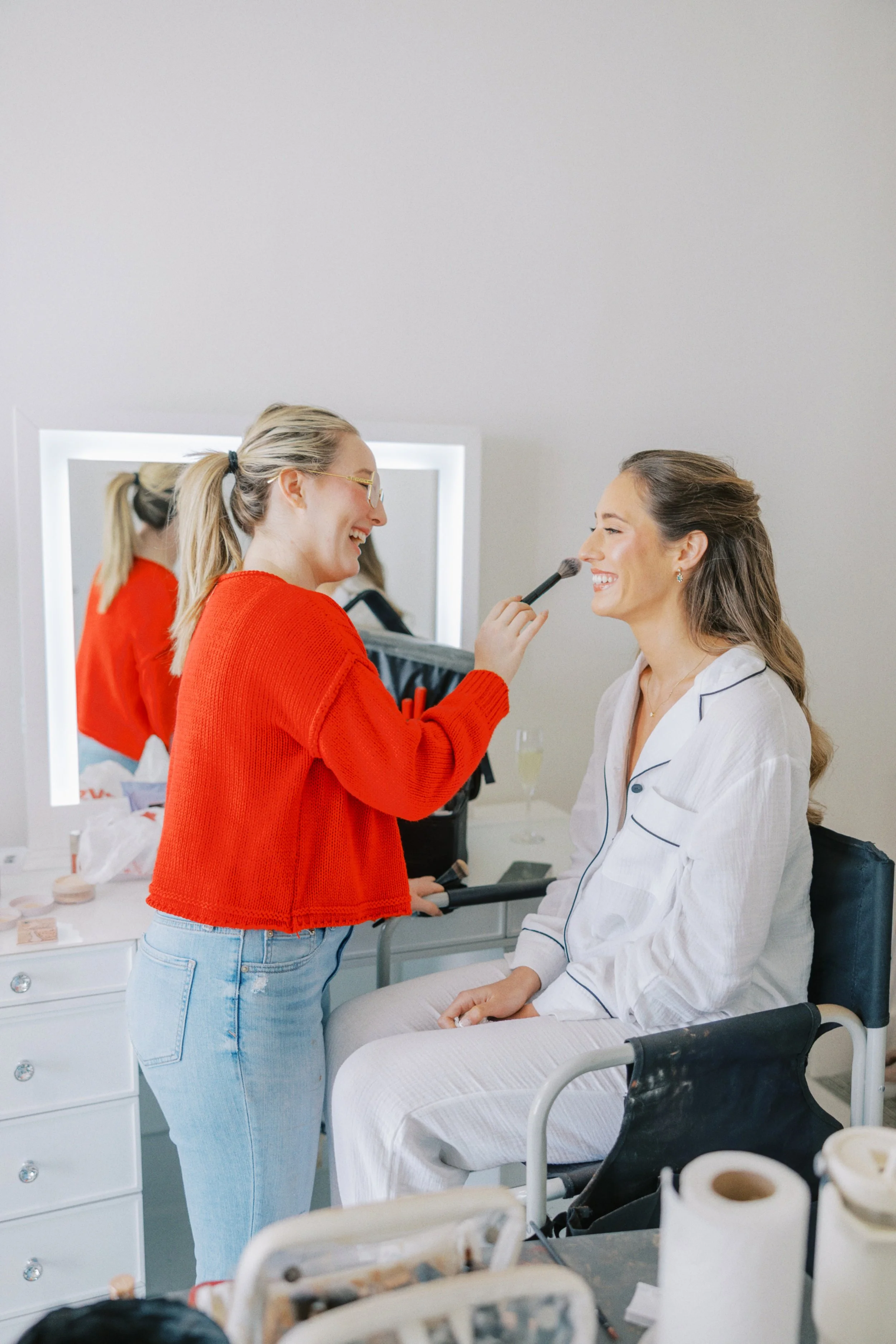 A makeup artist in a red sweater is applying makeup to a woman seated in a wheelchair, both smiling and enjoying the moment, in a bright room with a mirror in the background.