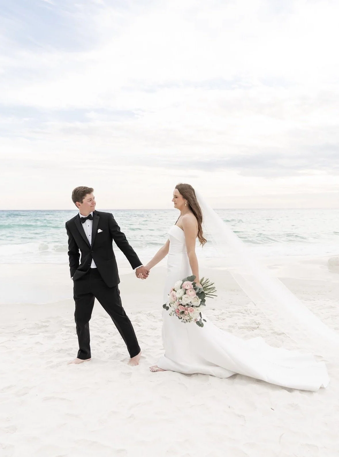 A bride and groom holding hands on a beach, with the ocean and sky in the background, dressed in wedding attire.