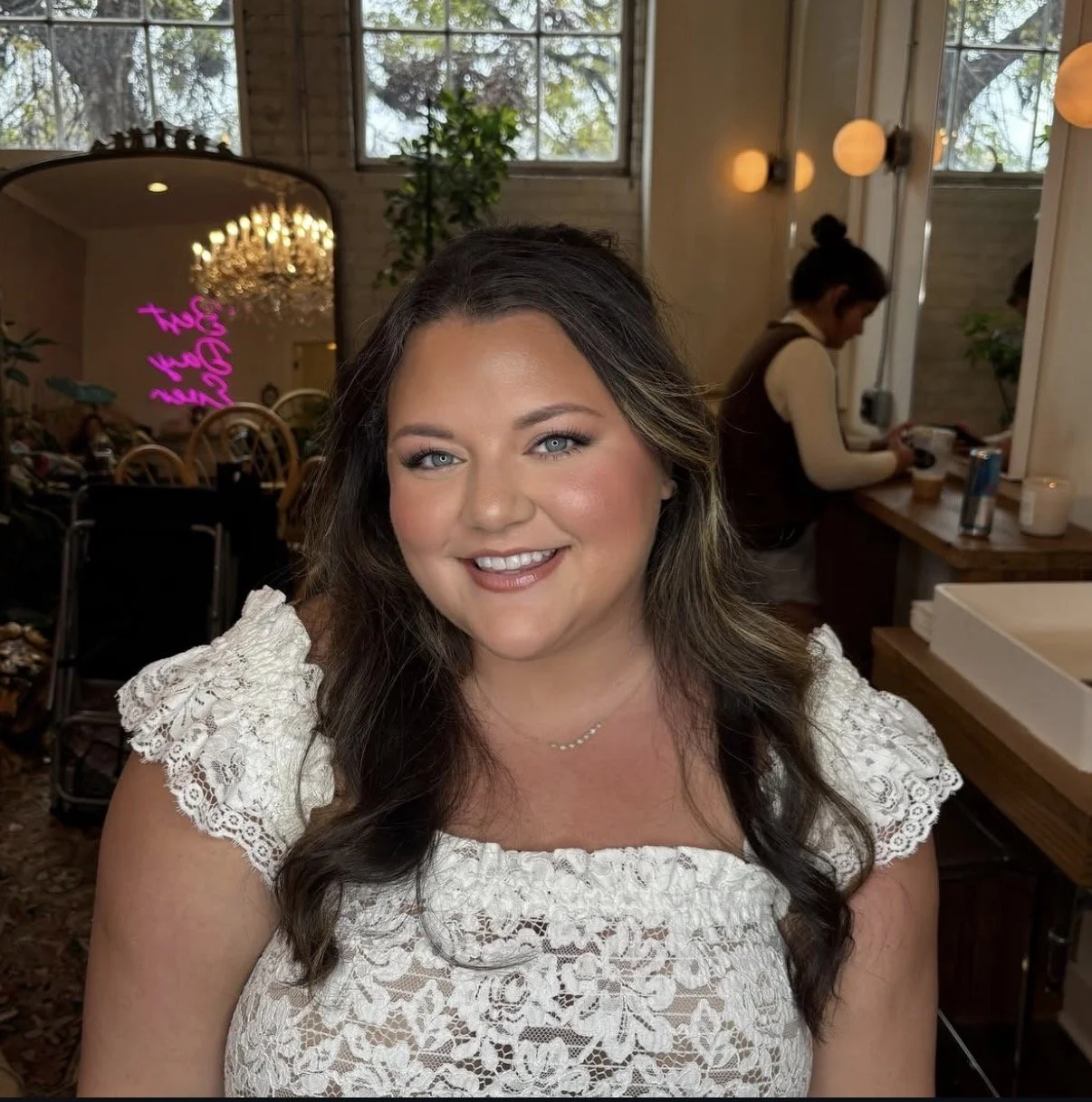 A smiling woman with wavy brown hair and blue eyes, wearing a white lace top and a delicate necklace, in a cozy cafe with warm lighting and a large window.
