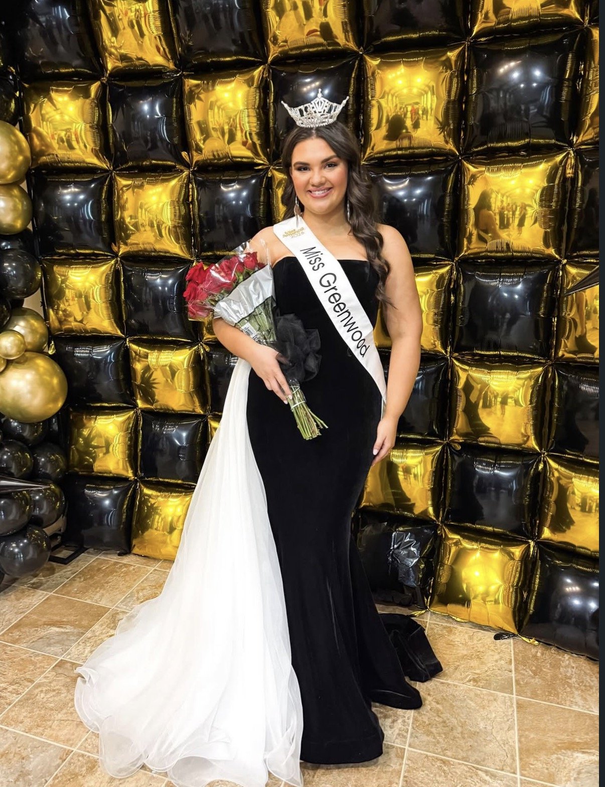 A young woman wearing a black and white gown, a crown, and a sash that says 'Miss Greenwood' is holding a bouquet of red roses. She is standing in front of a backdrop made of black and gold balloons.