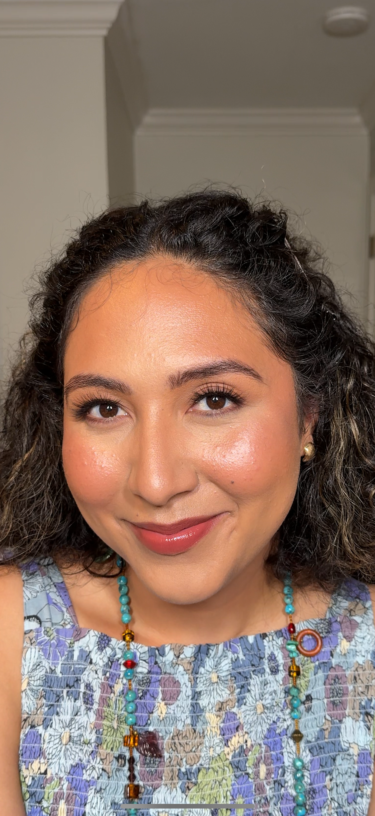A woman with curly dark hair, wearing a floral top, colorful beaded necklace, and earrings, smiling at the camera in a well-lit indoor setting.