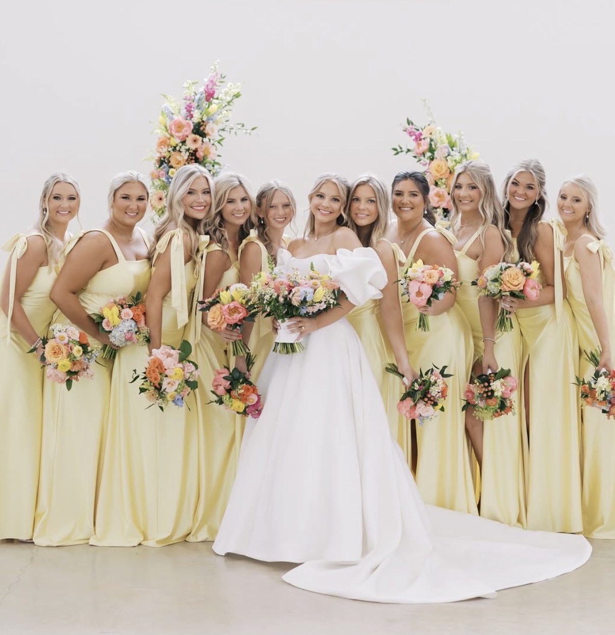 A bride in a white wedding dress standing in the center with ten bridesmaids in yellow dresses holding bouquets of pink, orange, and yellow flowers around her, against a plain white background with floral arrangements.