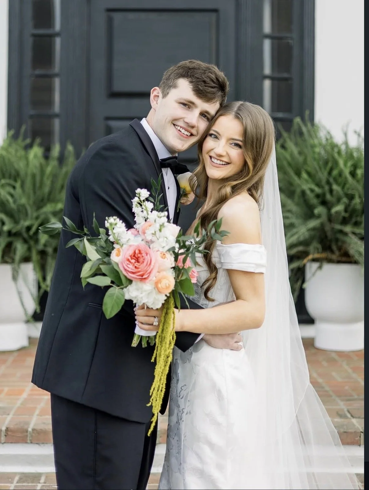 A newlywed couple smiling and embracing outside in front of a dark door and greenery. The groom is in a black tuxedo with a bowtie and the bride is in a white off-the-shoulder wedding dress holding a bouquet of pink and white flowers.