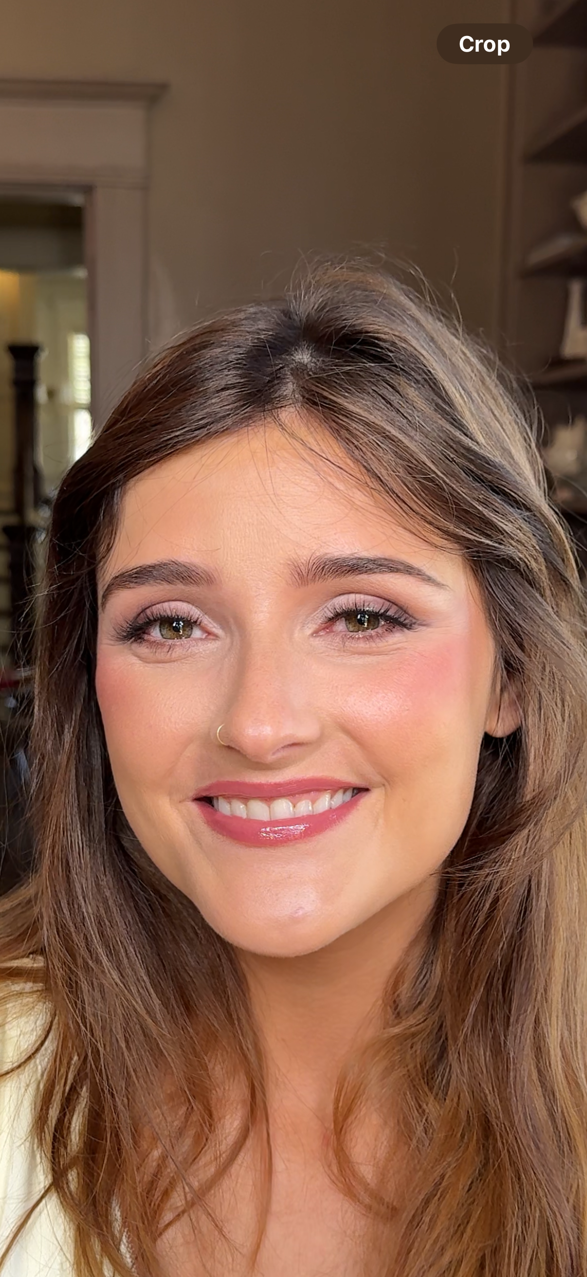 Close-up of a smiling woman with brown hair, light makeup, and a nose piercing in a well-lit indoor setting.