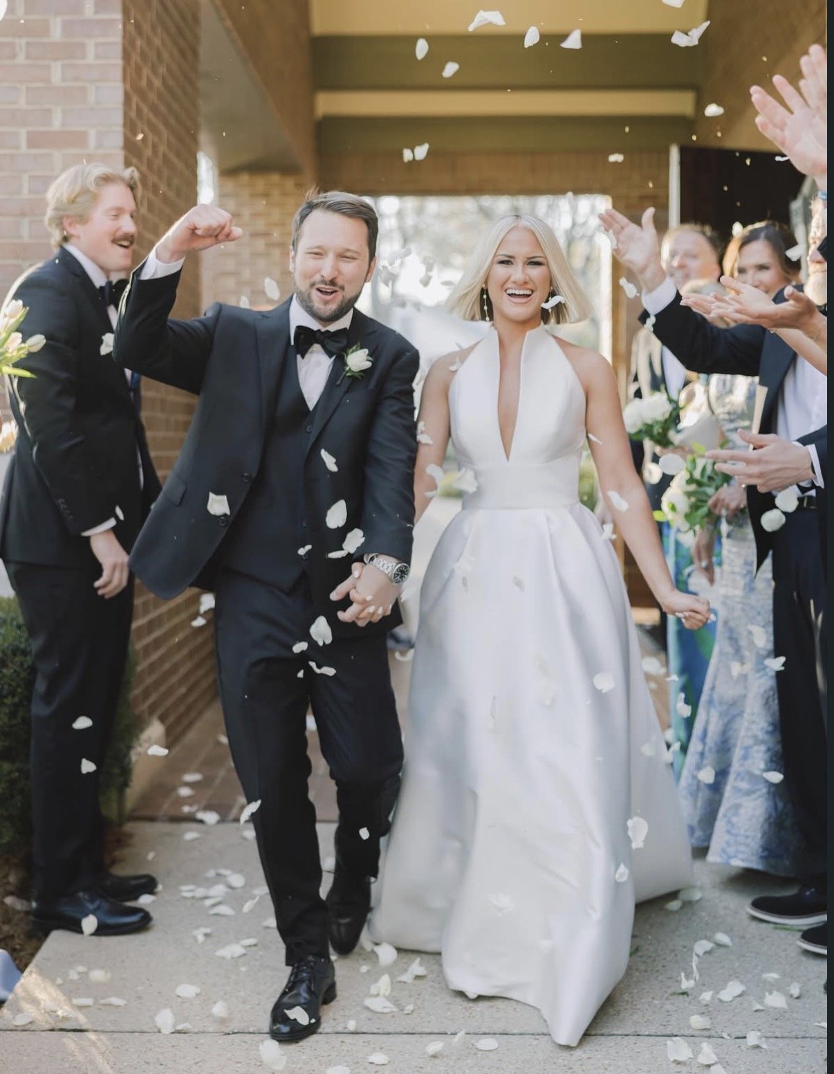 A newlywed couple walking through a shower of flower petals outside a building, holding hands and smiling. The groom is wearing a black tuxedo with a bow tie, and the bride is in a white wedding dress. Friends and family in formal attire are celebrat