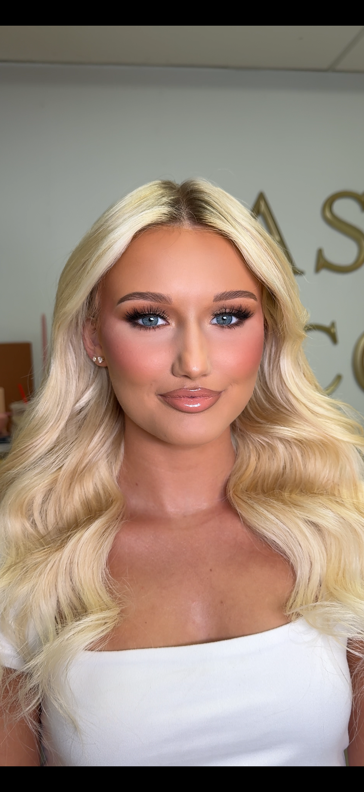 Close-up of a woman with long blonde wavy hair, blue eyes, makeup, and wearing pearl earrings, in front of a neutral background.