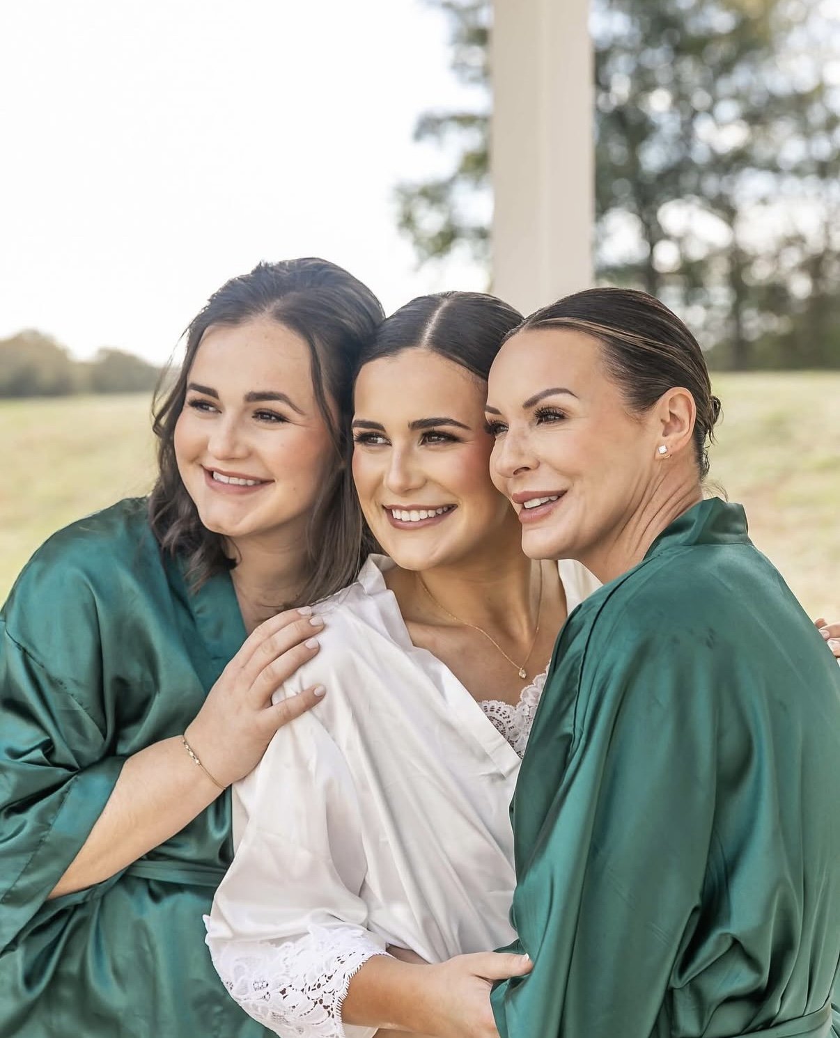Three women dressed in graduation robes are smiling and hugging each other outdoors.