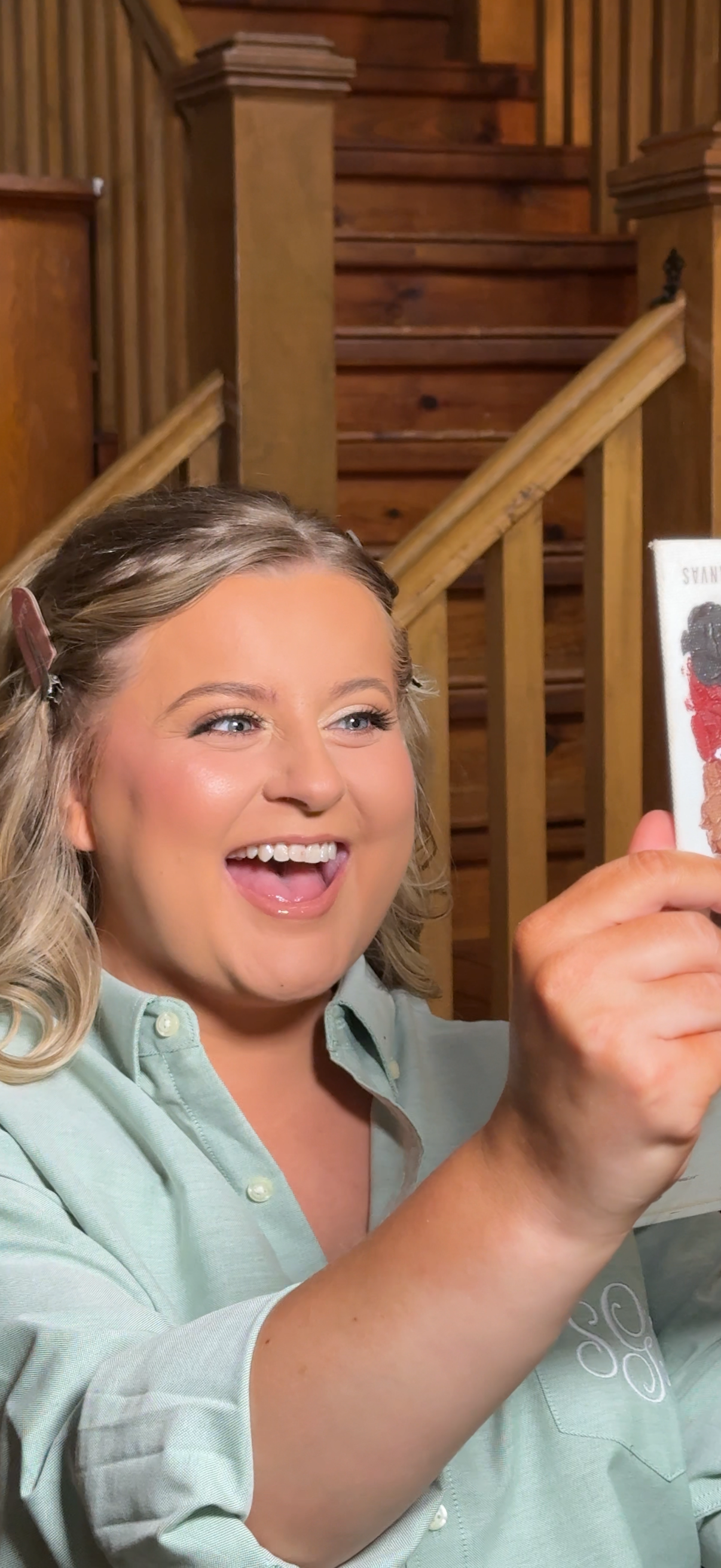 A woman with blonde hair smiling and holding a colorful card, standing in front of wooden stairs and railing.