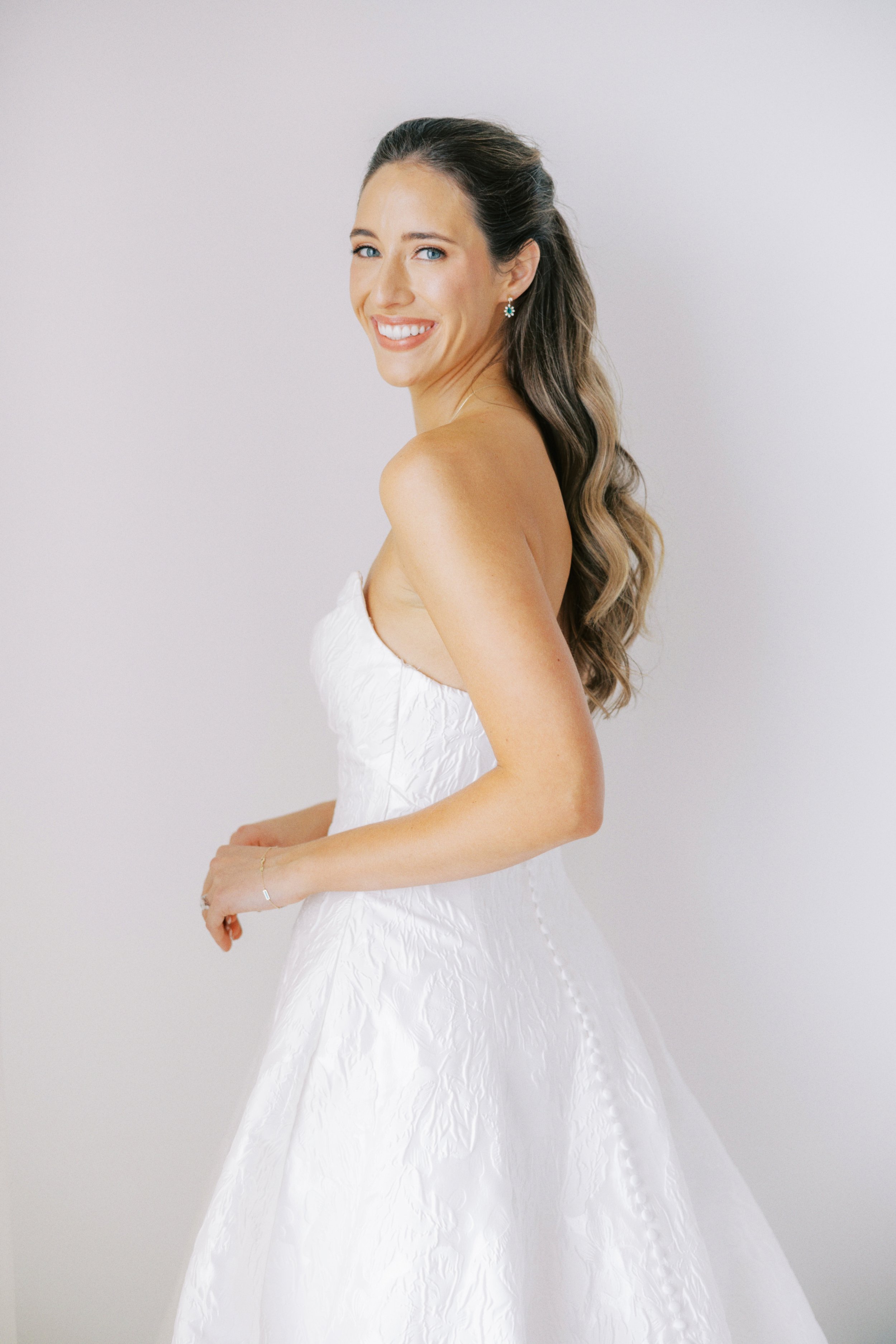 A woman in a strapless white wedding dress, with wavy brown hair, smiling and looking at the camera.
