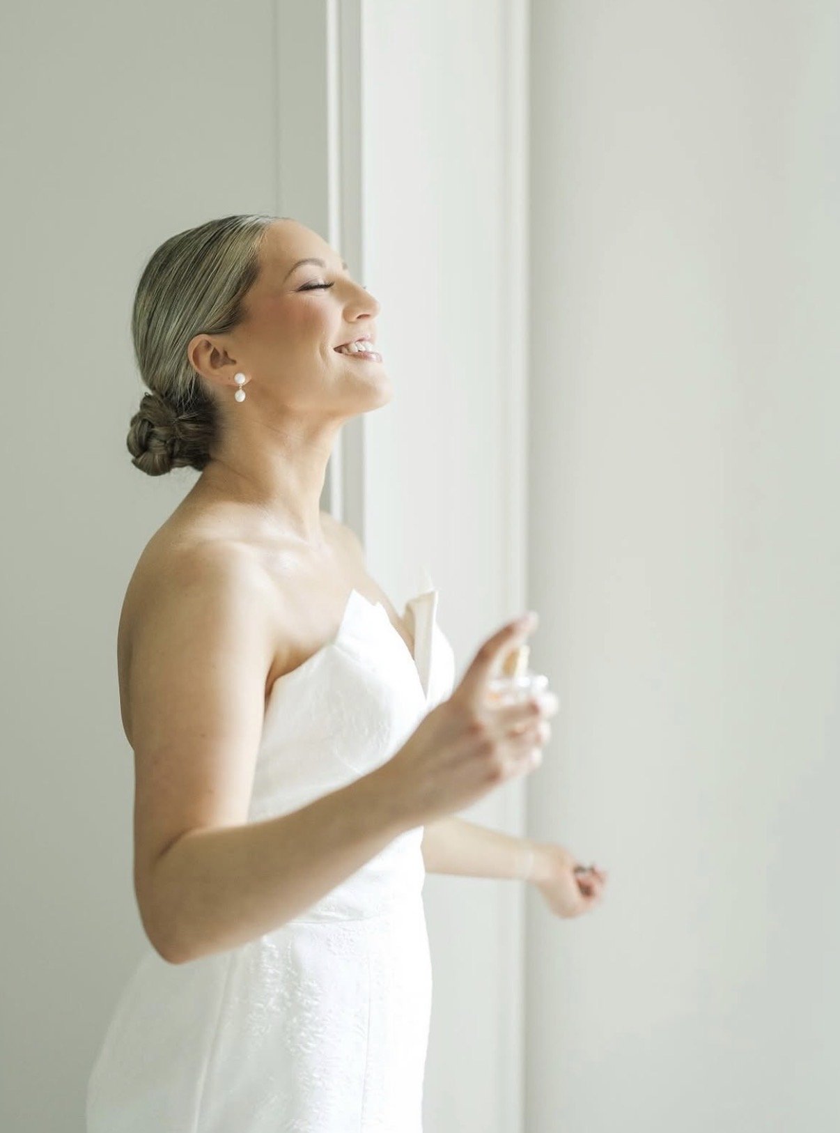 A woman in a white wedding dress with pearl earrings, smiling happily with her eyes closed, standing near a window with natural light.