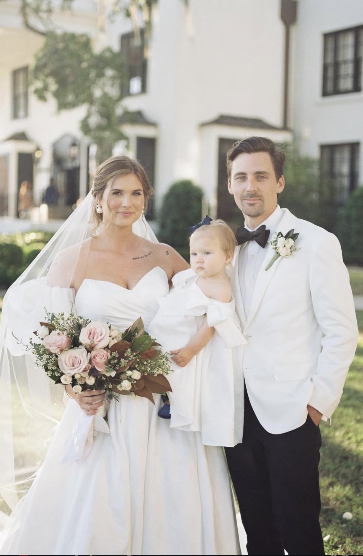 Wedding photo of a bride, groom, and young girl standing outdoors with white buildings in the background. The bride is holding a bouquet of pink and white flowers, wearing a white wedding dress and veil. The groom is dressed in a white tuxedo with a