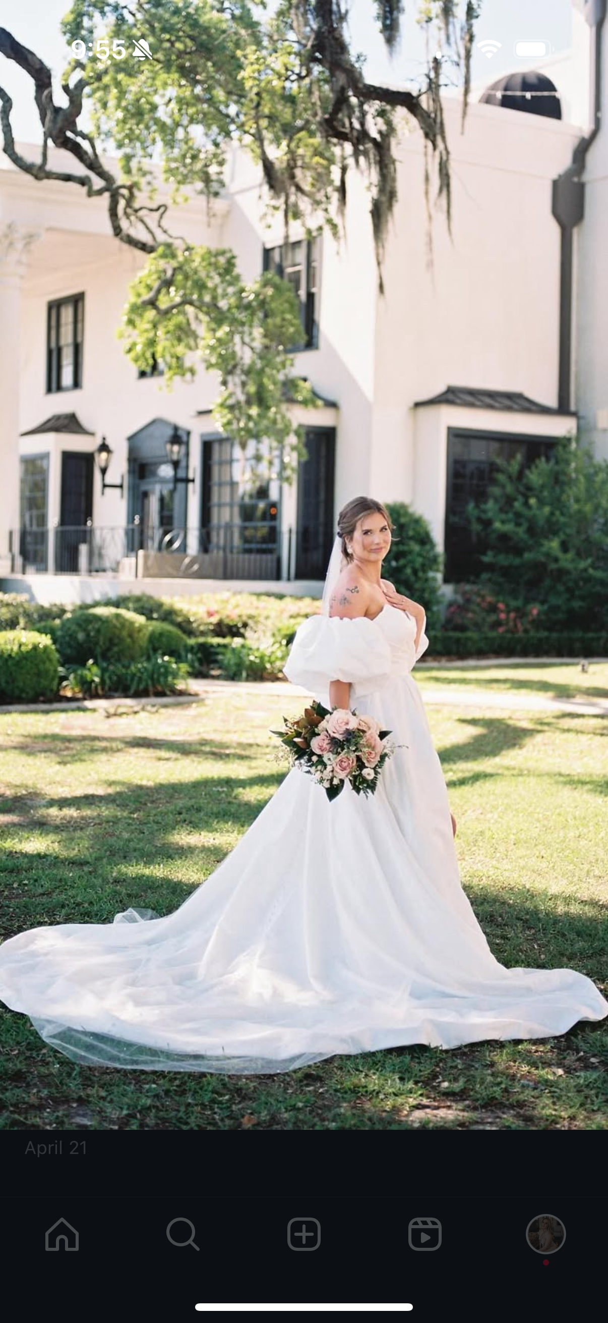 Bride in a white wedding dress holding a bouquet, standing on a grassy lawn in front of a white building with black-framed windows and doors, shaded by tree branches.